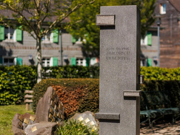 Graue Gedenkstele für Friedrich Ueberweg auf begrüntem Platz mit Bänken
