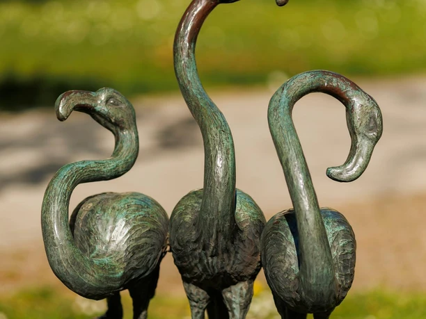 Drei bronzene Flamingo-Skulpturen auf einer Wiese mit unscharfem Parkhintergrund