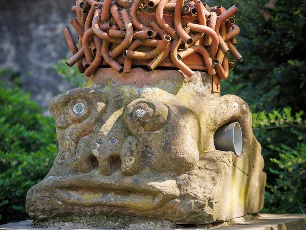 Skulptur „Musik im Kopf“ mit Röhrenhaar und Gesicht aus Stein im Grünen