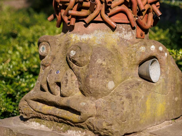 Skulptur „Musik im Kopf“ mit Steinkopf, rotem Röhrenhaar und Hörtrichter im Grünen