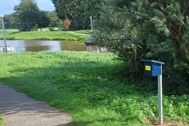 Du siehst eine Kurbelfähre am Fluss und eine blaue Stempelbox am Weg.