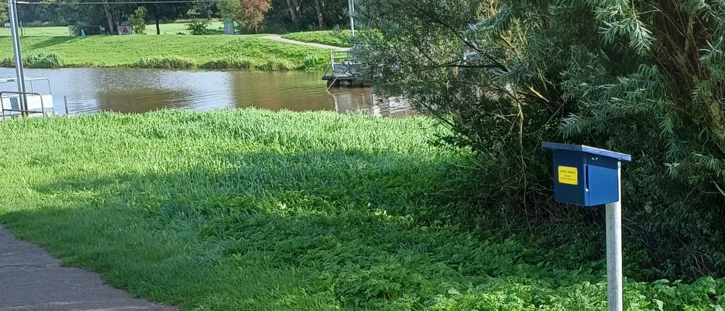 Du siehst eine Kurbelfähre am Fluss und eine blaue Stempelbox am Weg.
