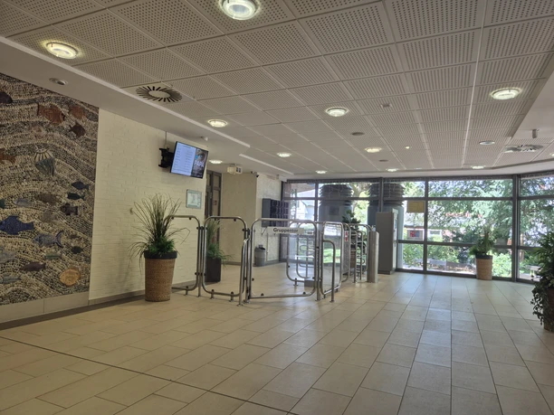 Eingangsbereich Hallenbad Foyer des Hallenbads mit Drehkreuzen, Pflanzen, Wandmosaik und großen Fenstern.