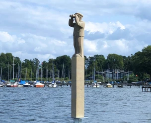 "Der Hafenmeister" Skulpturenpromenade Die Skulptur „Der Hafenmeister“ steht auf einem hohen Sockel am Wasser, umgeben von Segelbooten.