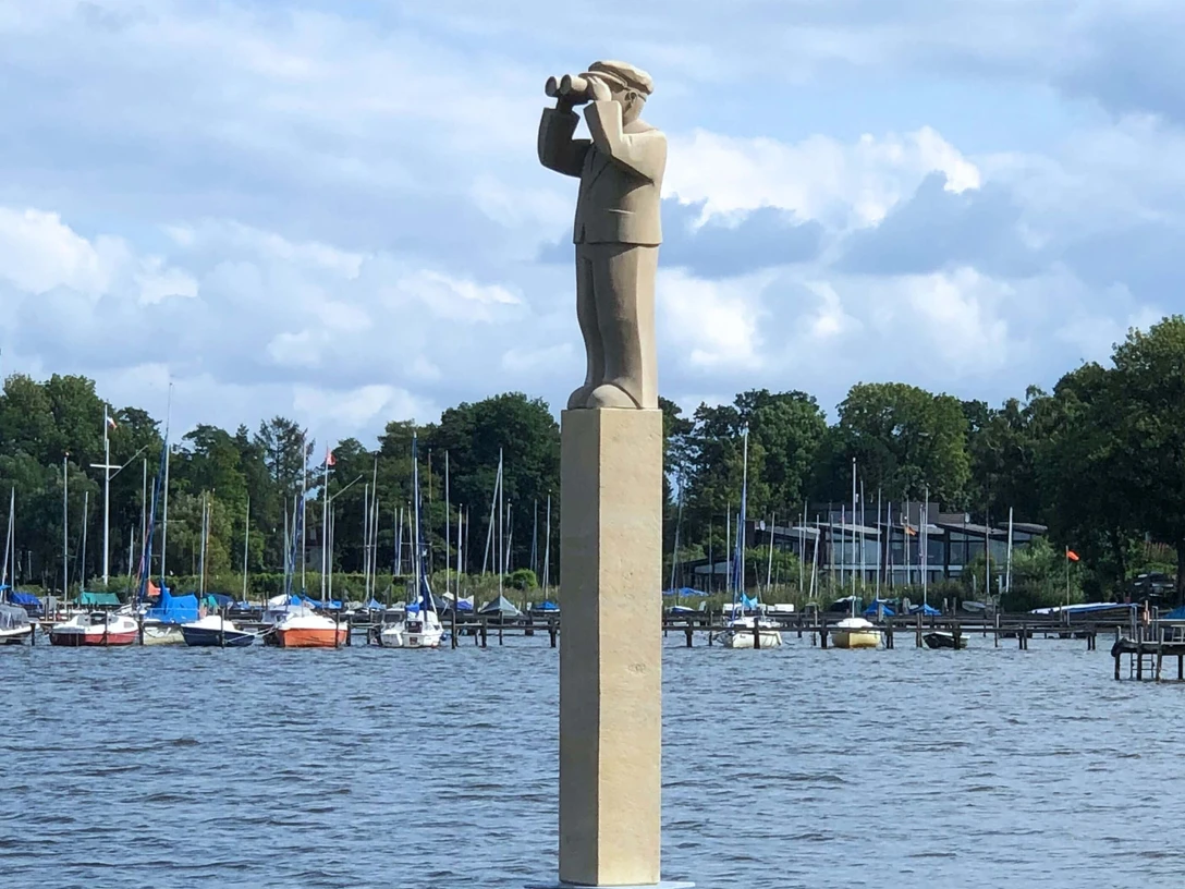 "Der Hafenmeister" Skulpturenpromenade Die Skulptur „Der Hafenmeister“ steht auf einem hohen Sockel am Wasser, umgeben von Segelbooten.The sculpture "The Harbour Master" stands on a high plinth by the water, surrounded by sailing boats.Skulpturen "The Harbour Master" står på en høj sokkel ved vandet, omgivet af sejlbåde.De sculptuur "The Harbour Master" staat op een hoge sokkel aan het water, omringd door zeilboten.