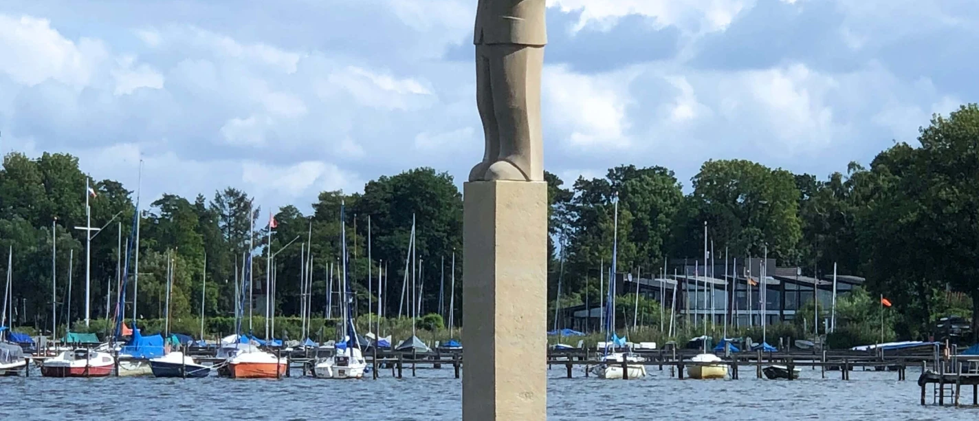 "Der Hafenmeister" Skulpturenpromenade Die Skulptur „Der Hafenmeister“ steht auf einem hohen Sockel am Wasser, umgeben von Segelbooten.