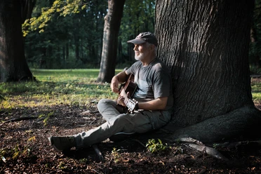 Gitarrist sitzt im Wald an einem Baumstamm und spielt im Sonnenlicht.