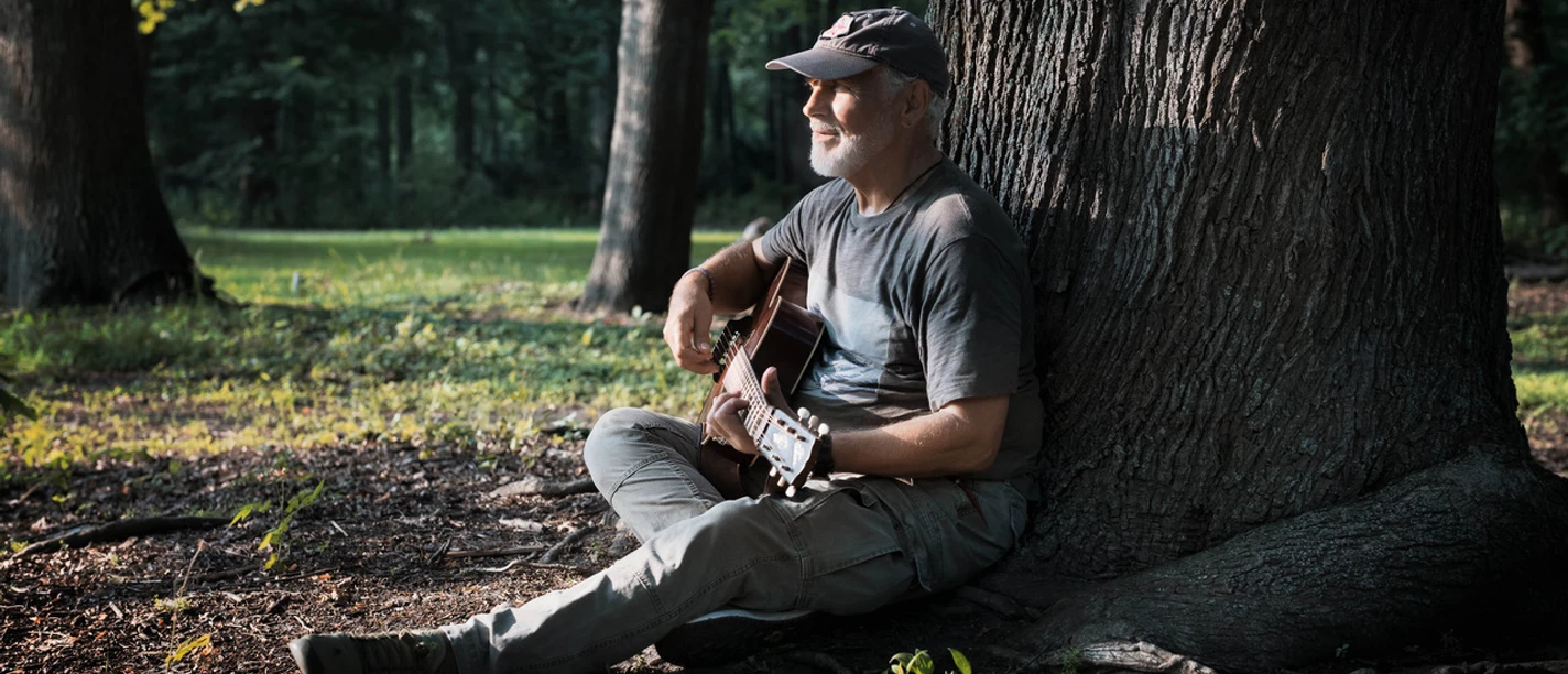 Gitarrist sitzt im Wald an einem Baumstamm und spielt im Sonnenlicht.