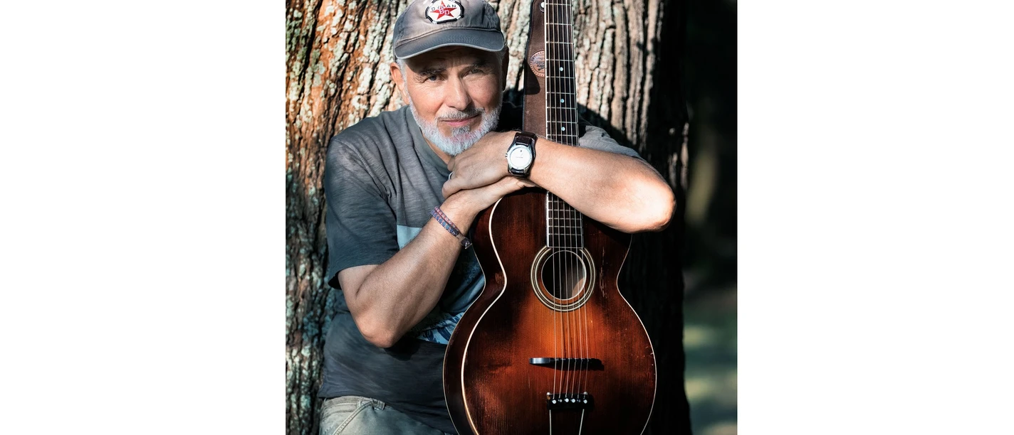 Hansa mit Akustikgitarre vor einem Baumstamm im Freien.