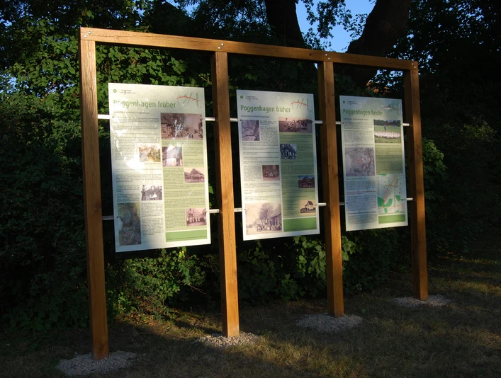 Dorftafeln Neu Drei Informationstafeln in Holzrahmen stehen auf einer Wiese vor dichtem grünem Laub im Abendlicht.Three information boards in wooden frames stand on a meadow in front of dense green foliage in the evening light.Tre informationstavler i trærammer står på en eng foran tæt, grønt løv i aftenlyset.Drie informatieborden in houten frames staan op een weiland voor dicht groen gebladerte in het avondlicht.