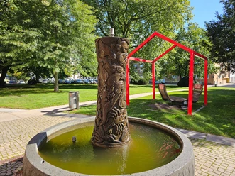 CDU-Brunnen mit Reliefsäule in rundem Becken, dahinter Parkbänke unter roten Rahmen.