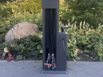 Fahrrad-Reparaturstation mit Werkzeugen und Luftpumpe vor begrüntem Beet.