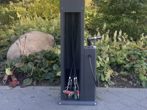 Fahrrad-Reparaturstation mit Werkzeugen und Luftpumpe vor begrüntem Beet.