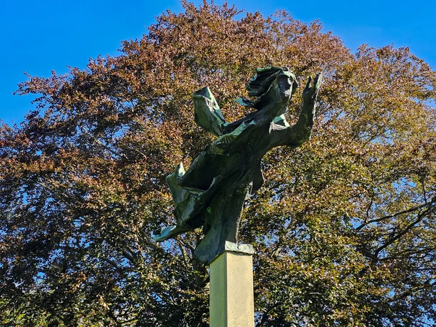 Bronzeskulptur Windsbraut auf hohem Sockel vor Baumkrone und blauem Himmel.