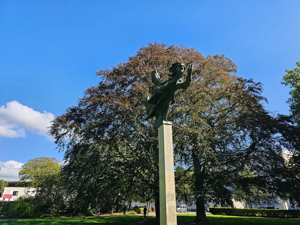 Skulptur Windsbraut auf hoher Stele vor großem Baum und blauem Himmel.