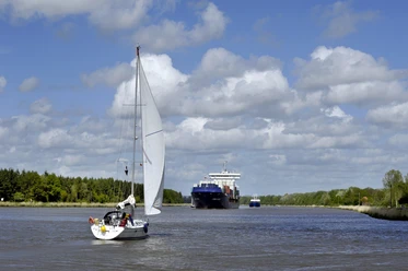 Große Pötte Segelboot und Frachter auf breitem Kanal.