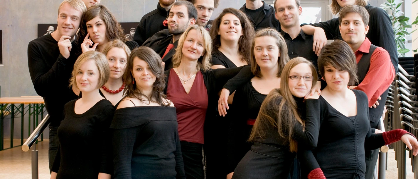 Eine Gruppe von 17 Personen steht zusammen und posiert für ein Foto. Sie tragen schwarze Kleider und lächeln freundlich in die Kamera.