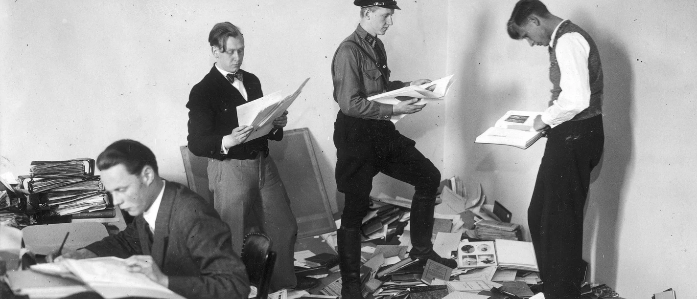 Bücher liegen chaotisch auf dem boden es sind 4 Männer zu sehen, 3 davon stehen (rechts) einer sitzt (links) und durchwühlen die Bücher