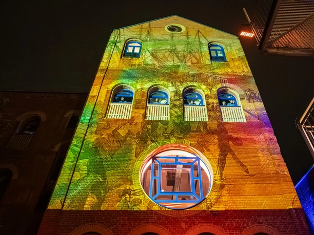 Bunte Lichtprojektion auf Backsteinfassade bei Nacht, Darstellende in Fenstern.