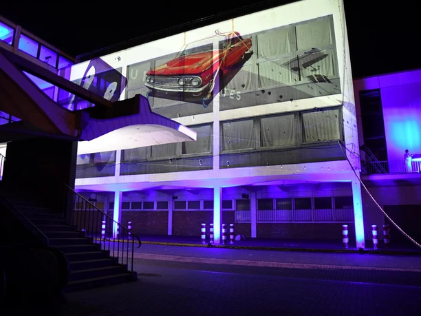 Nächtliche Fassadenprojektion auf einem Gebäude mit rotem Auto und violettem Licht.