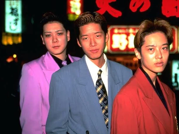 Filmstil zu "Shinjuku Boys" Drei junge Personen in bunten Anzügen vor Neonlichtern in Shinjuku