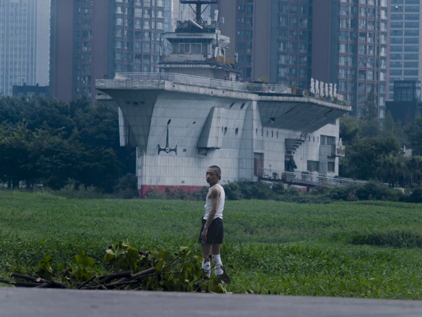 A person wearing a white top stands on a green meadow in front of a backdrop of urban high-rises and a concrete building