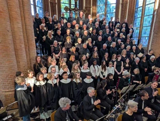 Großer Chor mit Kinderchor und Orchester vor hohen Fenstern einer Backsteinkirche.