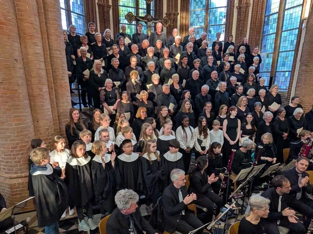 Großer Chor mit Kinderchor und Orchester vor hohen Fenstern einer Backsteinkirche.