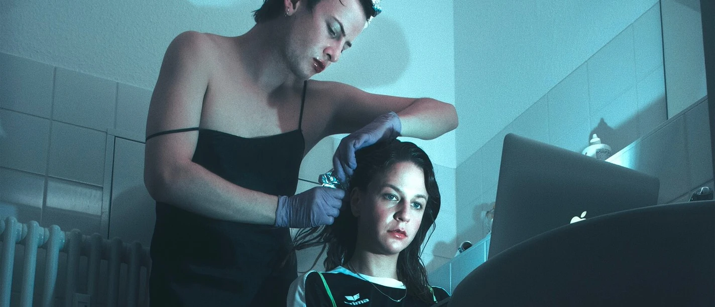 Two people in blue light; one is styling the other's hair in front of the sink.