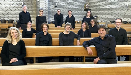 Gruppenfoto eines Orchesters in schwarzer Konzertkleidung, sitzend und stehend zwischen Kirchenbänken vor einer Steinwand.