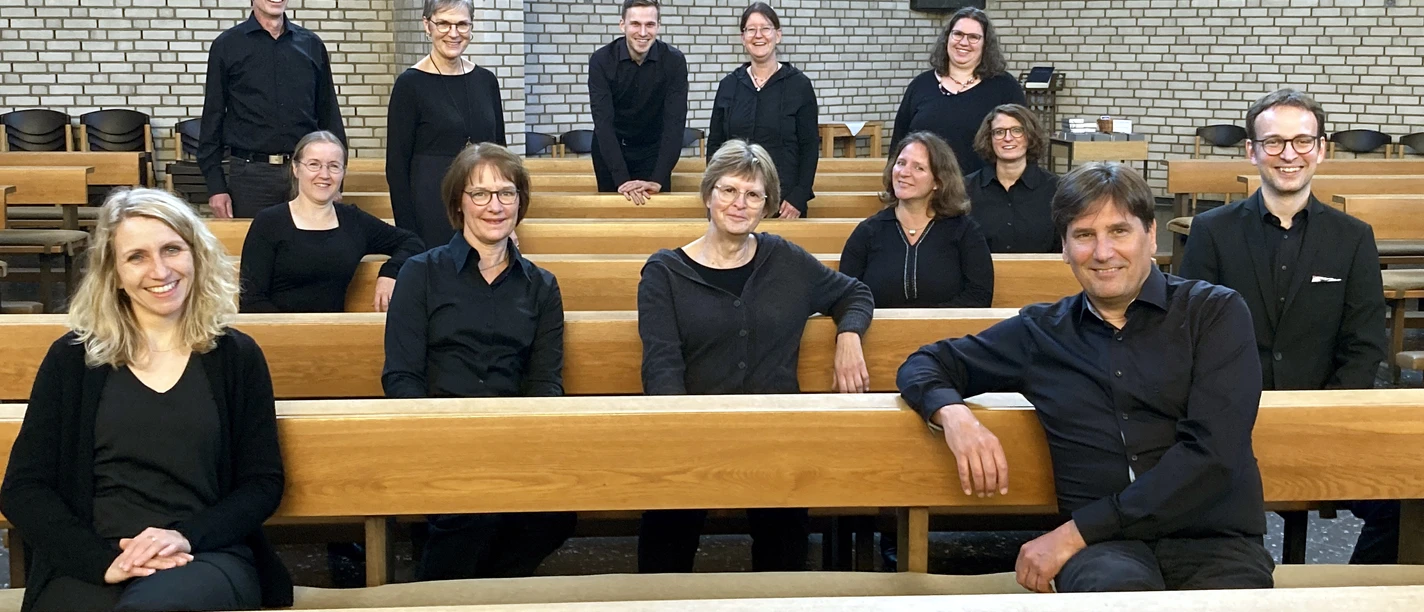 Gruppenfoto eines Orchesters in schwarzer Konzertkleidung, sitzend und stehend zwischen Kirchenbänken vor einer Steinwand.