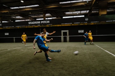 Traditions Cup at the BASE Street Soccer Arena A soccer player in blue shoots on the indoor field at Strassenkicker Base; opponents in yellow follow