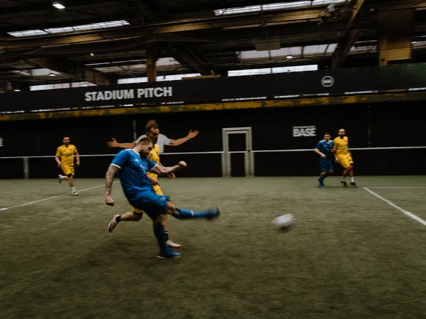 Traditions Cup at the BASE Street Soccer Arena A soccer player in blue shoots on the indoor field at Strassenkicker Base; opponents in yellow follow