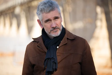 Curtis Stigers Ein Mann mit grauen Haaren steht in einem Außenbereich. Er trägt eine braune Jacke und einen schwarzen Schal.