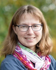 Eimler Eine lächelnde Frau mit Brille und einem bunten Schal. Im Hintergrund sind verschwommene grüne Pflanzen sichtbar.