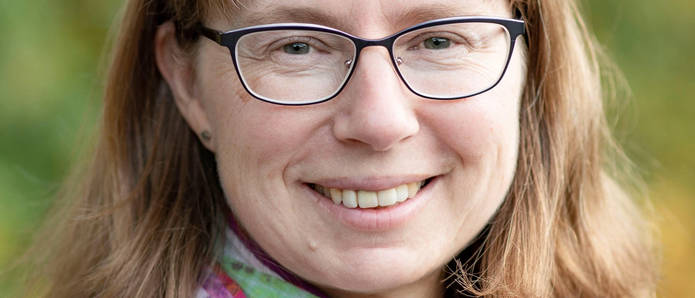 Eimler Eine lächelnde Frau mit Brille und einem bunten Schal. Im Hintergrund sind verschwommene grüne Pflanzen sichtbar.