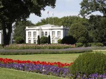 Das Haus des Gastes im Kurpark Weißes Gebäude im Kurpark umgeben von bunten Blumen, Grünflächen und hohen Bäumen im Hintergrund.