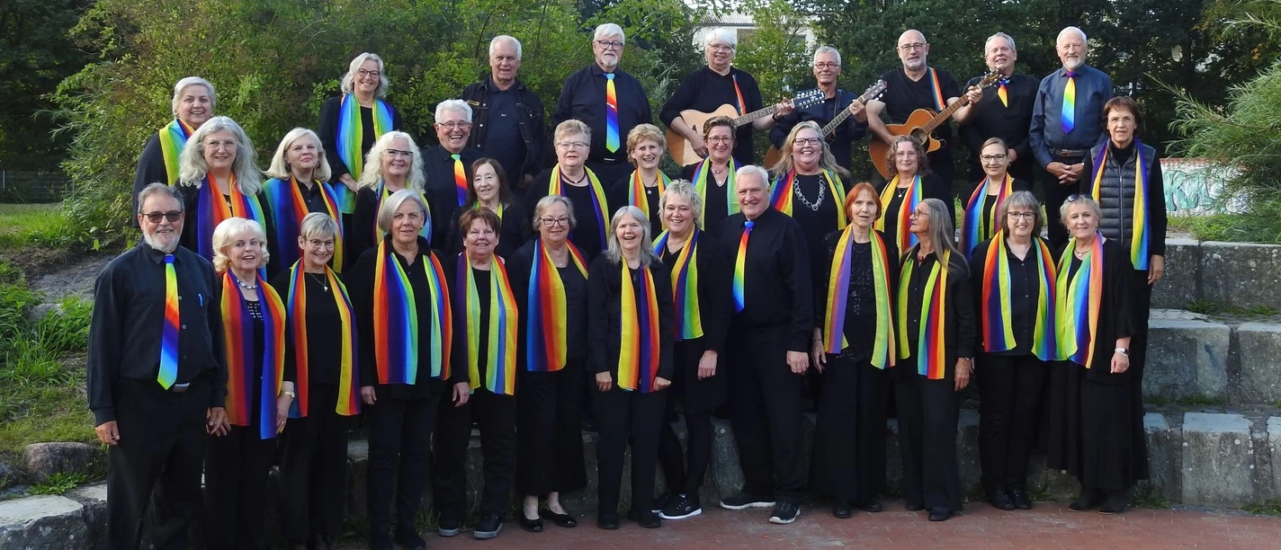 Chor Swinging Folk in Schwarz mit Regenbogenschals und Gitarren im Freien.