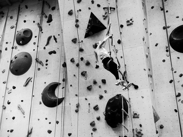 AHK Klettern Schwarz-Weiß-Aufnahme: Person klettert gesichert an hoher Indoor-Kletterwand.