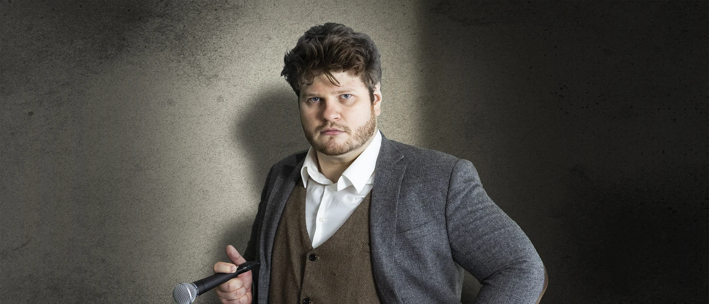 Jan Philipp Zymny mit Mikrofon und Requisitenpistole vor dunkler Wand.
