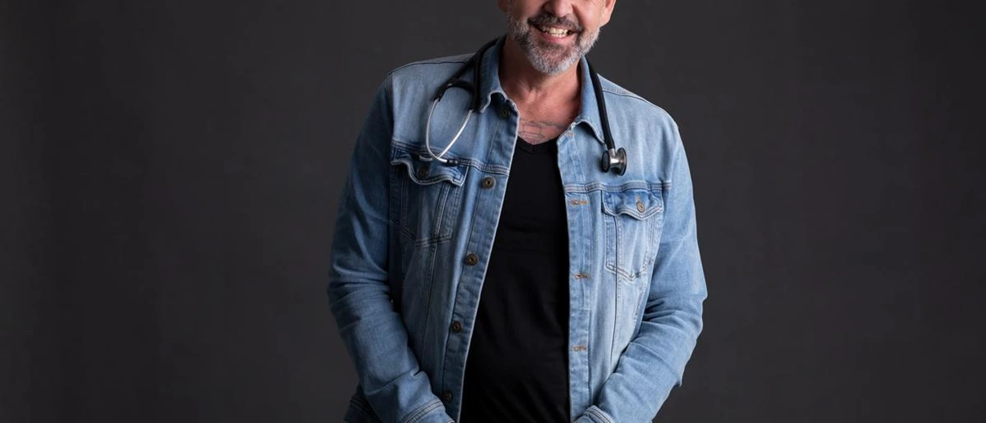 Ein lächelnder Mann trägt eine Jeansjacke und einen Hut. Er hat ein Stethoskop um den Hals und steht vor einem dunklen Hintergrund.