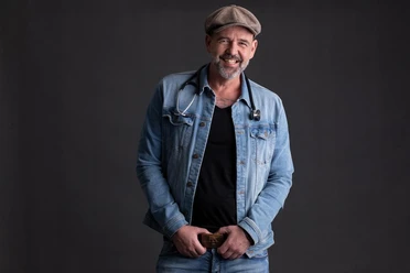 Doc Esser Ein lächelnder Mann trägt eine Jeansjacke und einen Hut. Er hat ein Stethoskop um den Hals und steht vor einem dunklen Hintergrund.