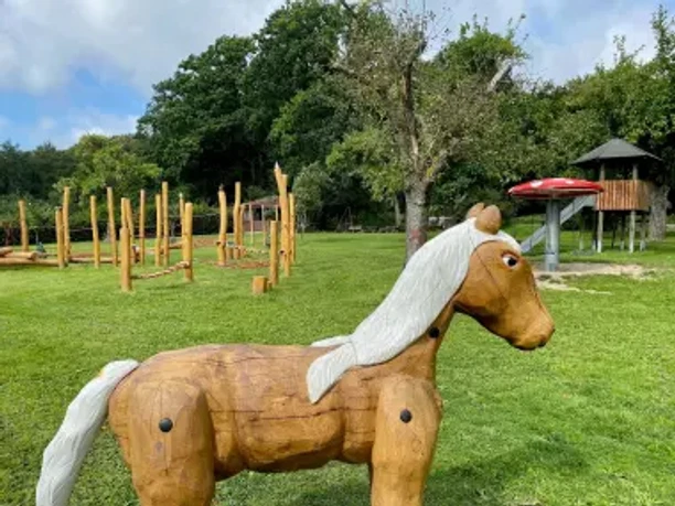 Spielplatz Freudeneck Geschnitztes Holzpferd auf einer Wiese mit Kinderspielplatz und Spielturm.