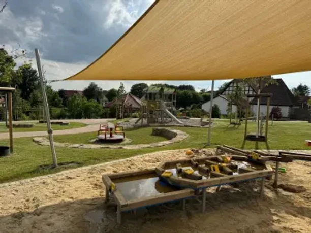 Mehrgenerationenpark Haldem Wasserspielstation im Sand unter einem Sonnensegel mit Spielplatzgeräten im Hintergrund.