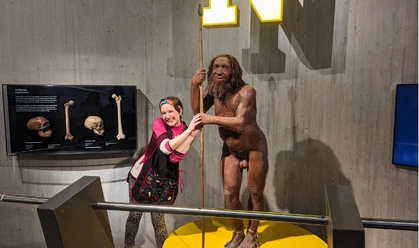 Comedyführung: "Mit dem Wischmopp durch die Steinzeit" Im Museum ist eine Putzfrau zu sehen. Sie steht sehr nah an einer lebensecht wirkenden Neandertaler-Figur.
