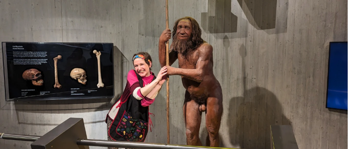 Comedyführung: "Mit dem Wischmopp durch die Steinzeit" Im Museum ist eine Putzfrau zu sehen. Sie steht sehr nah an einer lebensecht wirkenden Neandertaler-Figur.