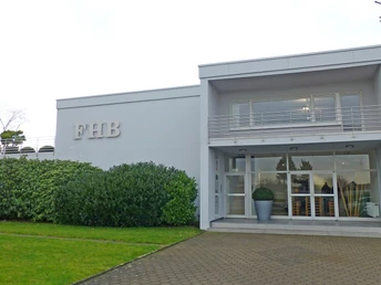 Die Firma FHB Berufsbekleidung Der Eingang der FHB Berufsbekleidung mit moderner Fassade, großen Fenstern und gepflegtem Grünbereich.