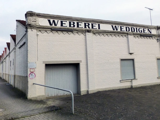 Die Leinenweberei Weddigen Historische Weberei mit weißer Fassade und Schriftzug "Weberei Weddigen", gepflasterter Hofzugang.