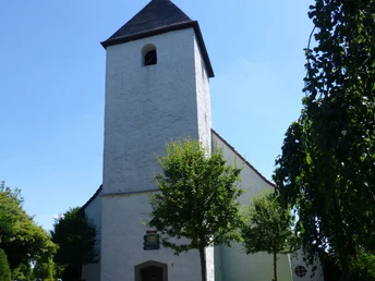 Die Autobahnkirche Exter Weiße Kirche mit hohem Turm und Wetterhahn, umgeben von Bäumen unter klarem, blauem Himmel.