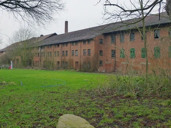Das Gut Deesberg Altes Backsteingebäude mit grünen Fensterläden, umgeben von grüner Wiese und kahlen Bäumen.